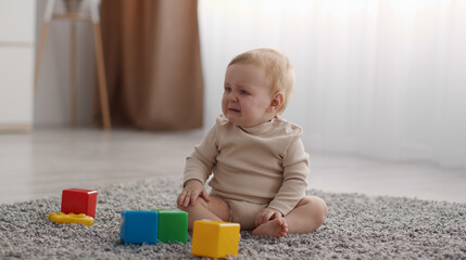 Portrait of cute little baby crying, sitting on floor with colorful cubes, suffering from pain or loneliness, free space