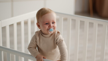 Sedative for toddlers. Little baby with pacifier standing in crib, looking aside at bedroom,...