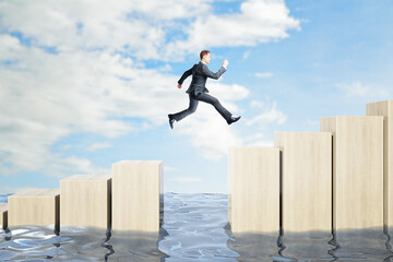 Businessman jumping over obstacles between wooden chart bars above water on bright blue sky...