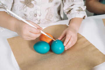 Cropped portrait of girl hand, painting multi coloring Easter hens eggs on brown paper plate table with paintbrush. Making art design white patterns on blue, red animal eggs. Holiday event. Close up