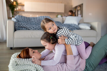 Little children lying on her mother and having fun in living room at home.