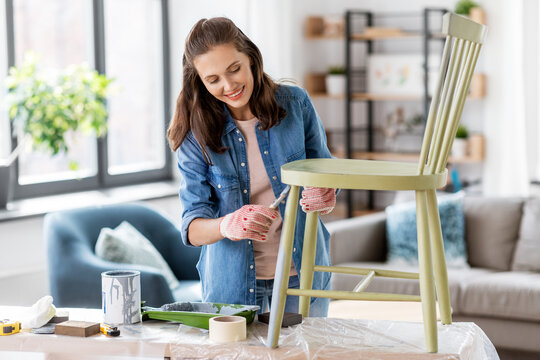 renovation, diy and home improvement concept - happy smiling woman in gloves with paint brush painting old wooden chair in grey color
