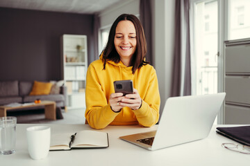 Smiling female freelancer using smart phone while sitting with laptop at home office