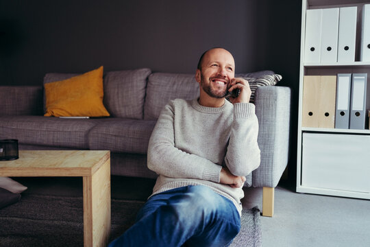 Smiling man sitting against sofa talking on mobile phone in living room at home