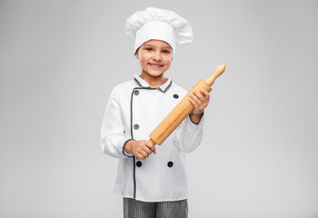 cooking, culinary and profession concept - happy smiling little girl in chef's toque and jacket with rolling pin over grey background
