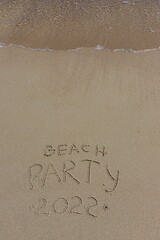 Fototapeta premium The inscription beach party 2022 on the sand by the water and the rising wave, seashore beach vacation by the sea