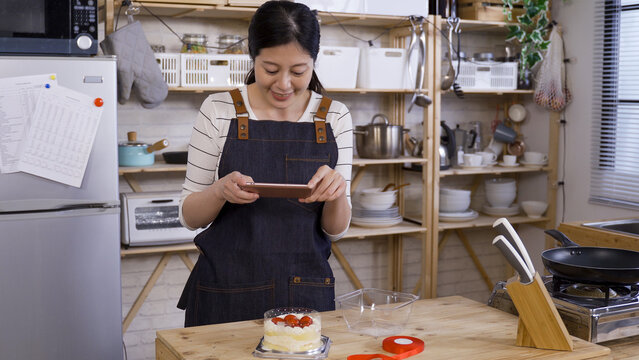 cheerful asian young girl running a small home baking business by kitchen table and using phone to take pictures for her product in a wooden style kitchen