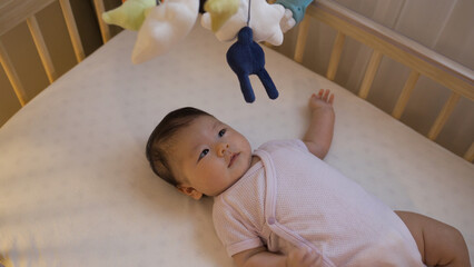 cropped adorable female infant in bodysuit is lying alone in the baby cot and looking at the...