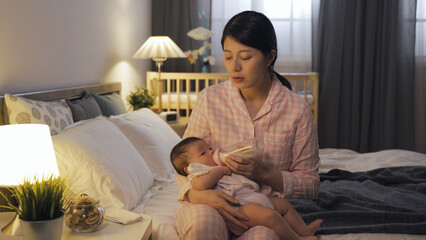 tired new mother sitting by lamp is dozing off while bottle feeding her newborn child at midnight...