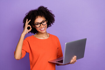 Photo of impressed young brunette lady hold laptop okey wear eyewear orange t-shirt isolated on...