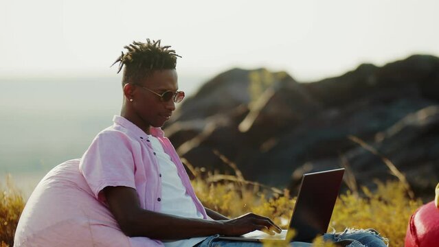 Black man sitting with laptop on chair bag in outdoor and drink coffee, side view. Black student freelancer is sitting in nature on the background of hills with a laptop. Concept education