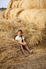 Heap of hay on field with little girl sitting in it smiling with sly pensive eyes looking away holding hay in hand wearing sundress. Having fun away from city on field full of golden hay.