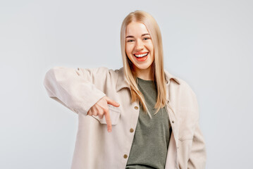 Portrait of smiling stylish blond girl in trendy shirt pointing finger down, showing promo deal, inviting to register, advertisement, standing over gray background
