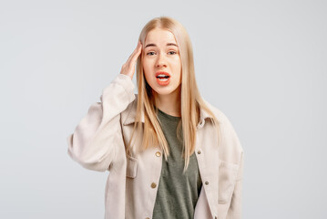 cute fair-haired young woman is thinking and holding hand on temple, recalling or remembering, wears casual clothes, stands against grey background. Body language concept