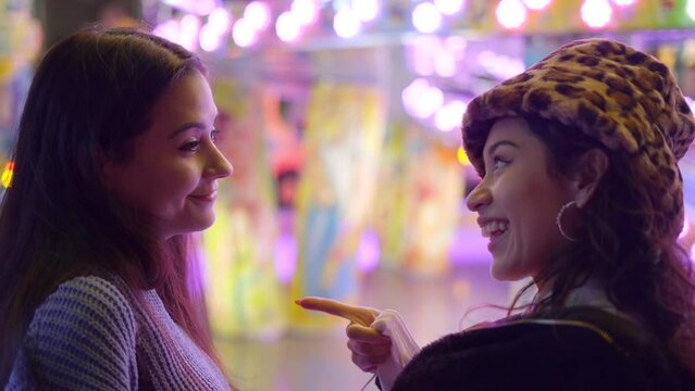 Two beautiful girls looking at each other nodding with happy face expression, agree on riding attraction at Valencia funfair at night