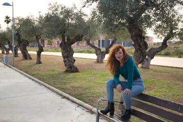 Young woman with red hair, freckles, sitting on a wooden bench smiling in an outdoor park. Concept happiness, joy, laughter, smile.