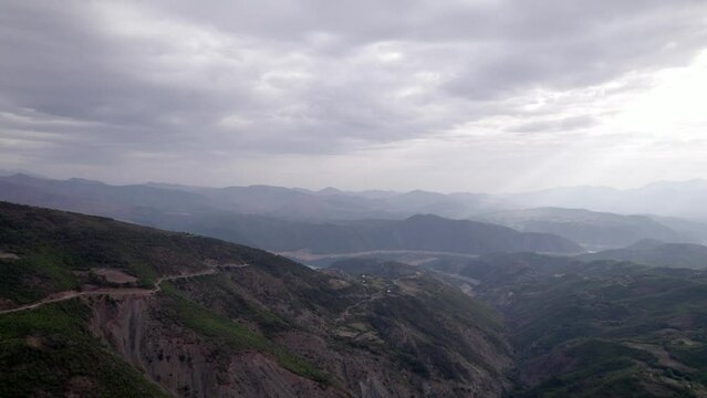 Frontal drone video ascending in "golden hour" over the mountains of SH22 in albania, over the village of Va Spas and in the background Lake Drin. cloudy sky letting the sun's rays through.