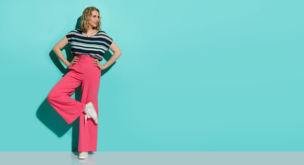 Stylish woman in pink loose pants, sneakers and striped blouse is posing on one leg and looking to the side.