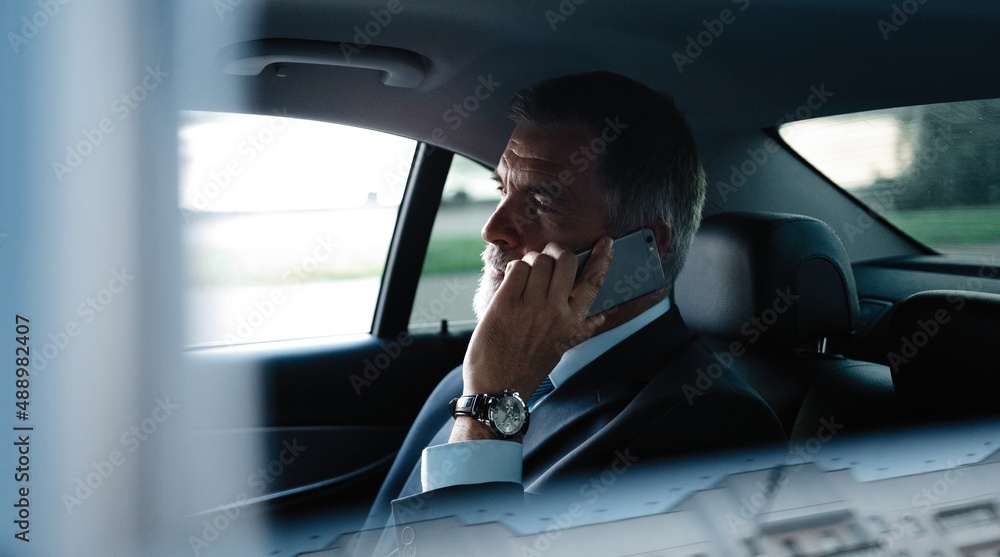 Wall mural Mature smiling man talking over phone while sitting in taxi. Businessman talking over cellphone while commuting.