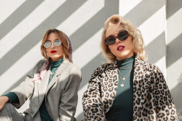 Two pretty young models girls with vintage sunglasses in trendy stylish clothes with fashionable leopard coat, green sweater and gray suit sits on the street near a wall with sunlight.