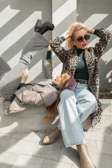 Two glamorous stylish girlfriends women in fashion trendy clothes with leopard coat, suit, jeans and shoes are sitting and posing in the city in the sunlight © alones
