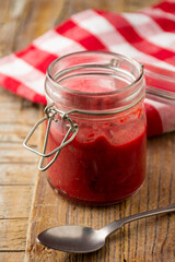 Top view of glass jar with homemade tomato and beet ketchup, on wood with red kitchen cloth and spoon, vertical