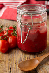 Close-up of glass jar with homemade tomato and beet ketchup, on wood with red kitchen cloth, cherry tomatoes and spoon, vertical