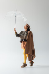 full length view of asian woman in leather pullover and yellow tights under transparent umbrella on grey