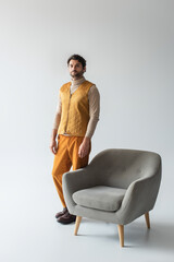 Fototapeta na wymiar full length view of man in yellow trousers and vest standing near armchair on grey