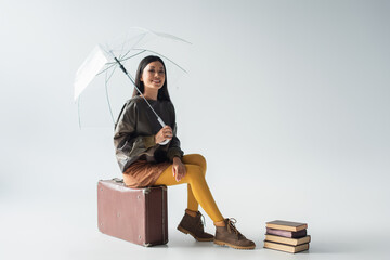 happy asian woman with transparent umbrella sitting on vintage suitcase near books on grey