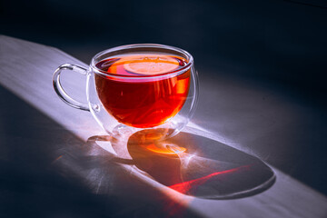 Close up of tea with lemon in transparent cup and sun glare on table. Hot drink of sparkling orange liquid in double glass cup with hard shadows and sunlight reflection, shine in line of sun light.