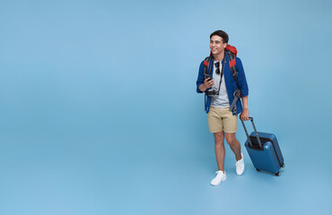 Happy young Asian tourist man holding smartphone with baggage going to travel on holidays isolated on blue copy space background. - obrazy, fototapety, plakaty