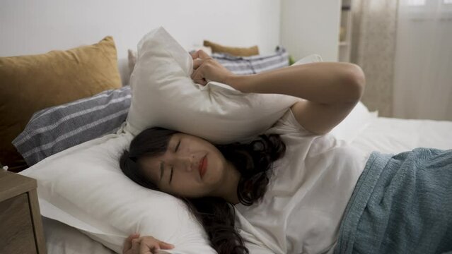 unhappy chinese girl annoyed with loud noise early is turning to cover her ears with hands and pillow trying to sleep on bed in the bedroom at home.