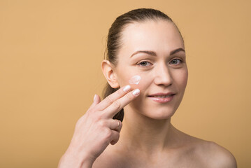 Portrait of a happy caucasian girl who applying a cosmetic cream on her face on her cheek isolated on a beige studio background. Skin care concept.