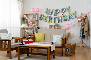 cozy home interior with birthday party decorations: pink balloons, birthday garland on white wall and gifts on living room table.