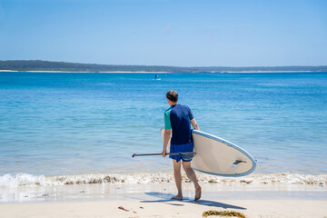 Man wearing rash guard with stand-up paddle board on a sandy beach near the ocean bay in Australia....