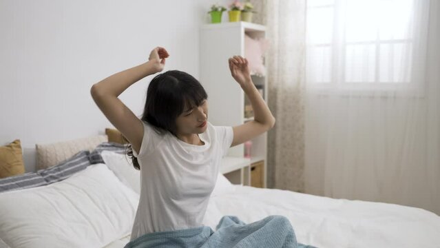 sleepy asian girl waking and rising from bed early in the morning is staring at the space after stretching up hands in the bedroom at home.