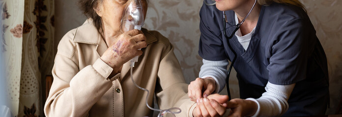 Young high-skilled pleasant woman doctor putting on nebulizer mask on face of her elderly female...
