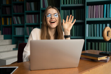 Workplace video communication manager uses laptop computer. Portrait of a female student with glasses in the office in the library.