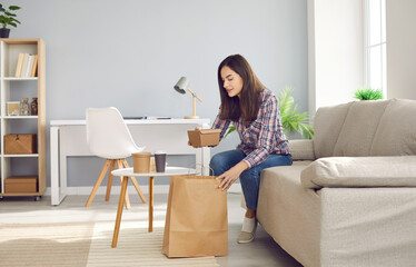 Happy beautiful woman opens paper bag with her takeaway meal. Young girl sitting on sofa at home...