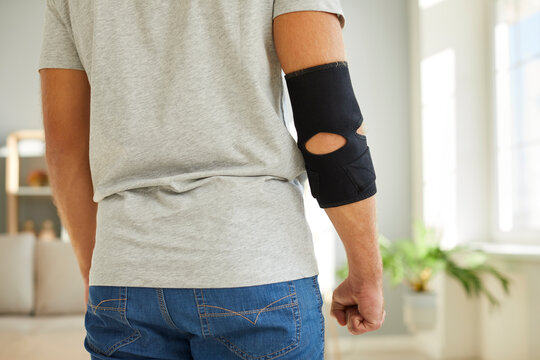 Young man wearing an adjustable black neoprene orthopedic elbow support brace on his right arm for easing pain in the elbow, back view, cropped close up shot. Concept of physical injury treatment