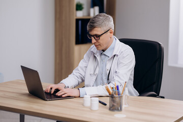 Male doctor wearing glasses and uniform working typing on computer. Family therapist consulting patients online through digital health platform.