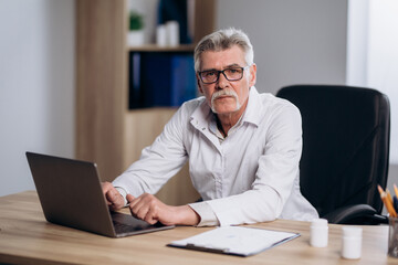 Serious male doctor sitting at workplace in hospital working on computer, looking thoughtful. Professional physician studying patient's electronic medical record.