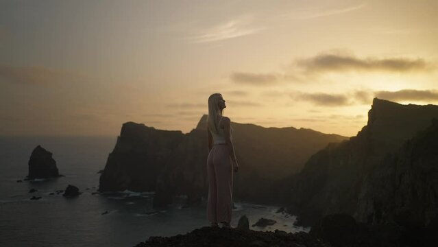 Blond traveler female in fashionable clothing enjoying dreamy sunrise, turns head, swaying hair, magical coastal viewpoint