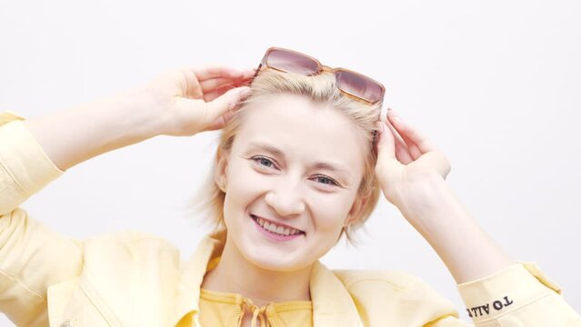 Young blond woman she takes off her sunglasses and looks to camera winking. Studio in light background