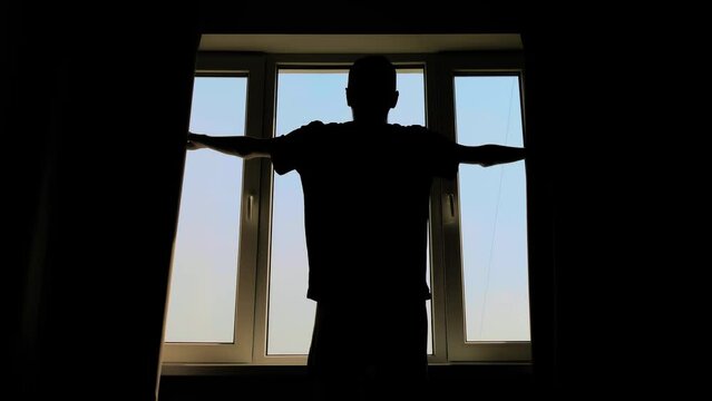 Silhouette of man opening curtains and looking out of window in room at morning - back view