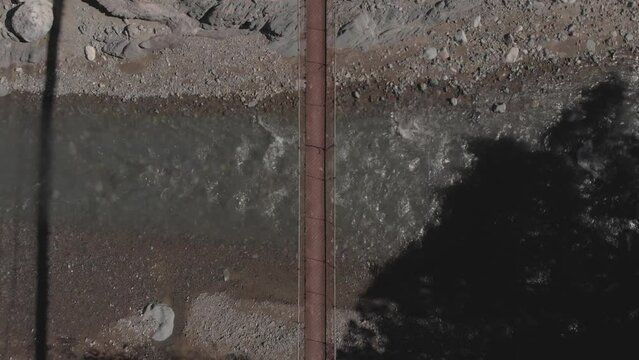 two people walking across a metal wire rope suspension bridge over rocky river water flowing down mountain valley Kabayan Benguet Philippines top down birds eye wide angle view