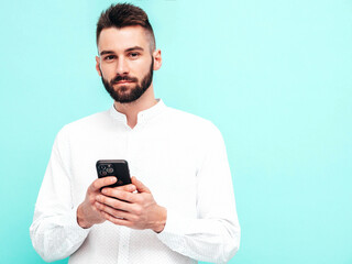 Handsome confident model.Sexy stylish man dressed in shirt and jeans. Fashion hipster male posing near blue wall in studio. Holding smartphone. Looking at cellphone screen. Using apps