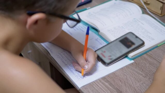 Schoolboy in Eyeglasses Writes with Pen, looking in Smart Phone. A left-handed child is sitting homework at the desk with books. Online a lesson with a gadget. Education concept. 4K. Close up.