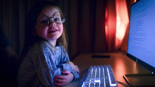 cute toddler baby girl in glasses, sitting at the compute in dark room